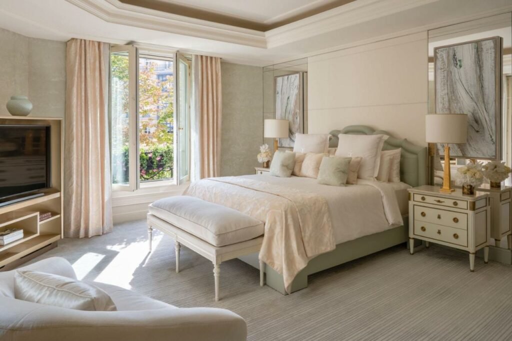 Quarto do Four Seasons Hotel George V Paris, um dos hotéis com café da manhã em Paris. Uma cama de casal está encostada na parede ao lado direito com mesinhas de cabeceira e abajures dos dois lados, assim como um banco aos pés. Logo em frente há uma estante com TV e uma poltrona.Na parede ao lado esquerdo da cama fica uma janela.