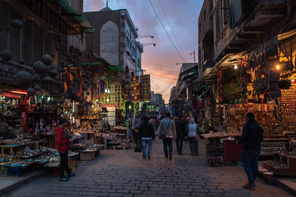 Uma movimentada rua do mercado de Khan El Khaliliao anoitecer, alinhada com barracas coloridas e iluminada por luzes penduradas. Pessoas são vistas caminhando e fazendo compras.