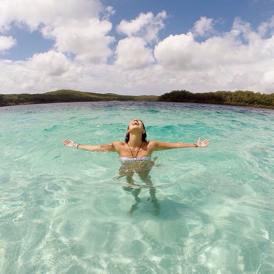 Uma pessoa em traje de banho está em águas azul-turquesa cristalinas em Fraser Island Austrália com os braços estendidos, olhando para um céu parcialmente nublado.