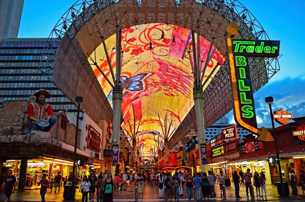 Teto de LED na Fremont Street. Ele se estende pela rua inteira e é todo colorido e iluminado, Diversas pessoas caminham abaixo dele. Do lado delas estão vários estabelecimentos.