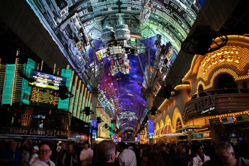 Teto de LED na Fremont Street. Ele se estende pela rua inteira e é todo colorido e iluminado, Diversas pessoas caminham abaixo dele. Do lado delas estão vários estabelecimentos, também cheios de luzes. A imagem ilustra o post de pontos turísticos em Las Vegas.