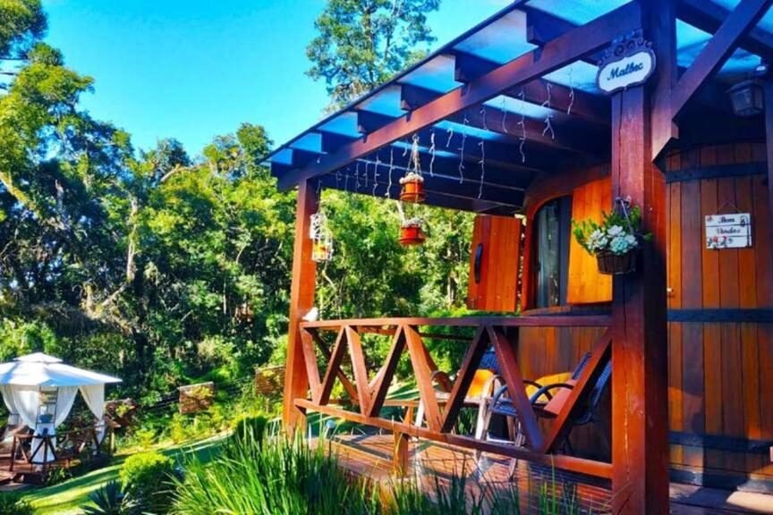 Foto do exterior do chalé Temático em Pipa de Vinho. A foto mostra uma cabana de madeira com uma varanda frontal proeminente, que possui um corrimão e uma estrutura de telhado. Na área gramada em frente à cabana, há cadeiras e mesas, sugerindo um espaço para sentar ou jantar ao ar livre. O ambiente ao redor da cabana é repleto de vegetação, árvores e um céu azul claro, indicando um cenário sereno e natural. Na varanda, há pequenas decorações penduradas e uma placa com a palavra “Chalet”