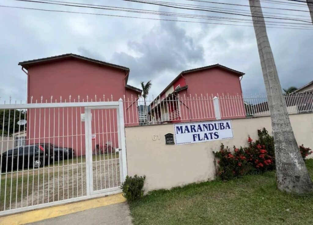 Imagem da frente do Flat em praia de Maranduba/Ubatuba com grama na calçada com um coqueiro do lado esquerdo da imagem, ao fundo um muro e atrás a propriedade.