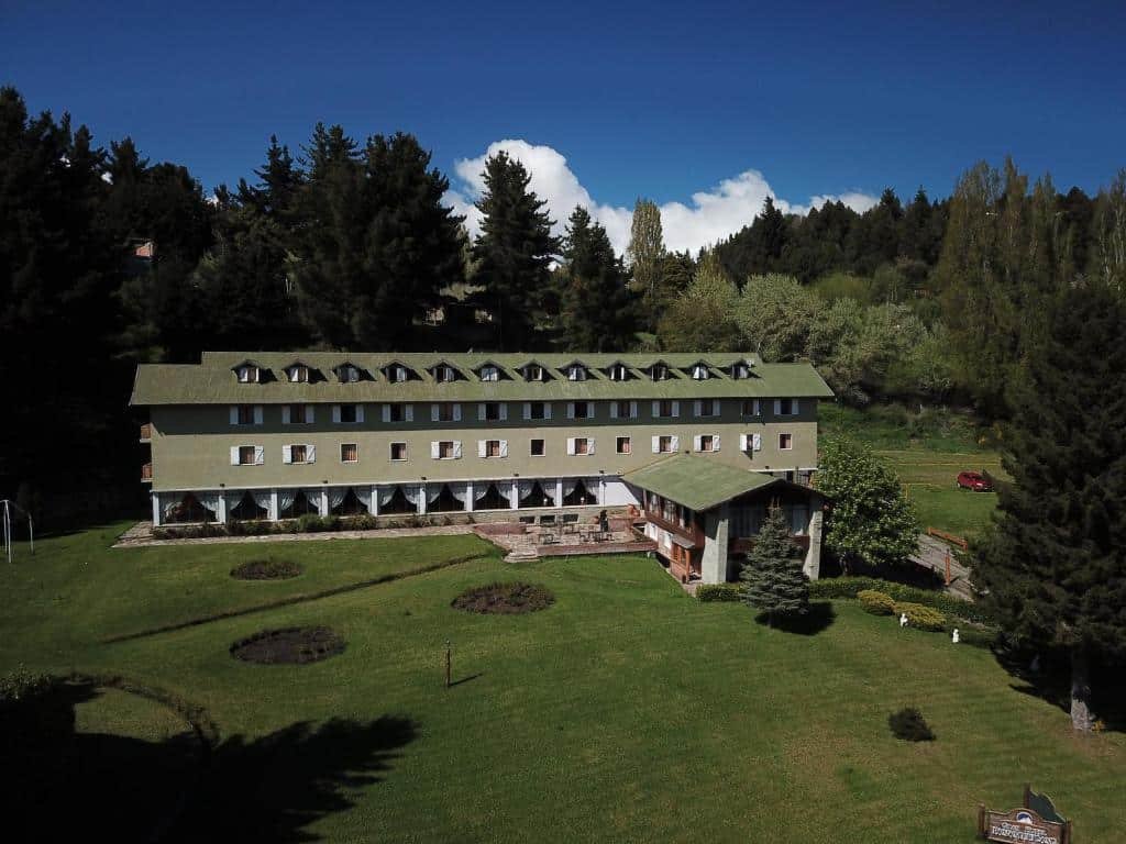 Vista da frente do Gran Hotel Panamericano com várias janelas de vidro, teto com telhados verdes, em volta da hospedagem tem bastante grama verde e ao fundo várias árvores. Representa hotéis em Bariloche