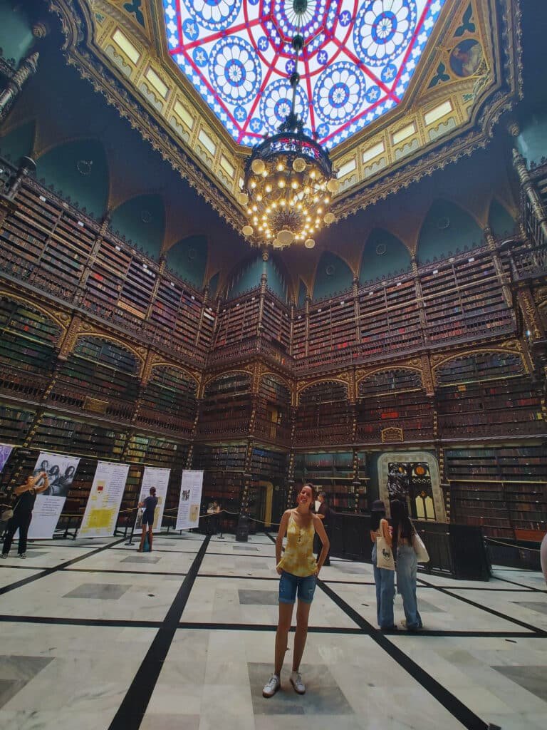 Uma mulher está em uma biblioteca ornamentada com um grande teto de vitral e prateleiras altas cheias de livros.