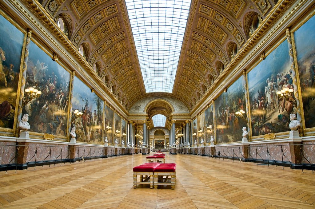 Corredor espaçoso e ornamentado com pinturas clássicas, bancos vermelhos, bustos em pedestais e um grande teto com claraboia.