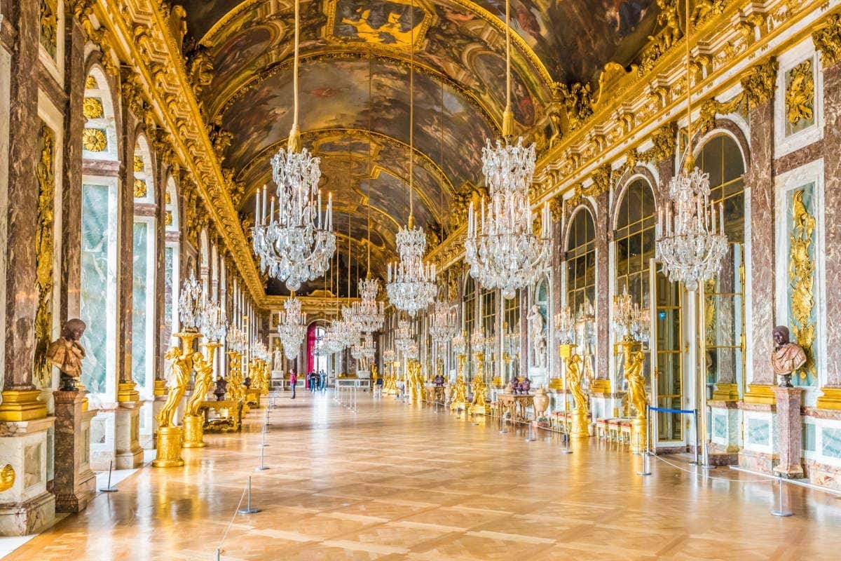 Um grande salão com lustres ornamentados, espelhos arqueados e um teto pintado no Palácio de Versalhes. Paredes e esculturas têm detalhes dourados.