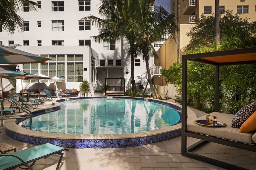A imagem mostra uma área de piscina circular cercada por espreguiçadeiras e guarda-sóis coloridos no Generator Miami, um dos hotéis em Miami Beach indicados. A piscina é ladeada por um edifício branco de vários andares, com grandes janelas. À direita, há uma cama de dia coberta por uma estrutura de madeira escura com um dossel laranja, cercada por vegetação tropical. Uma mesa ao lado da cama de dia tem um drink colorido e um petisco. O ambiente é tranquilo e acolhedor, ideal para relaxar em meio a palmeiras e plantas verdes.