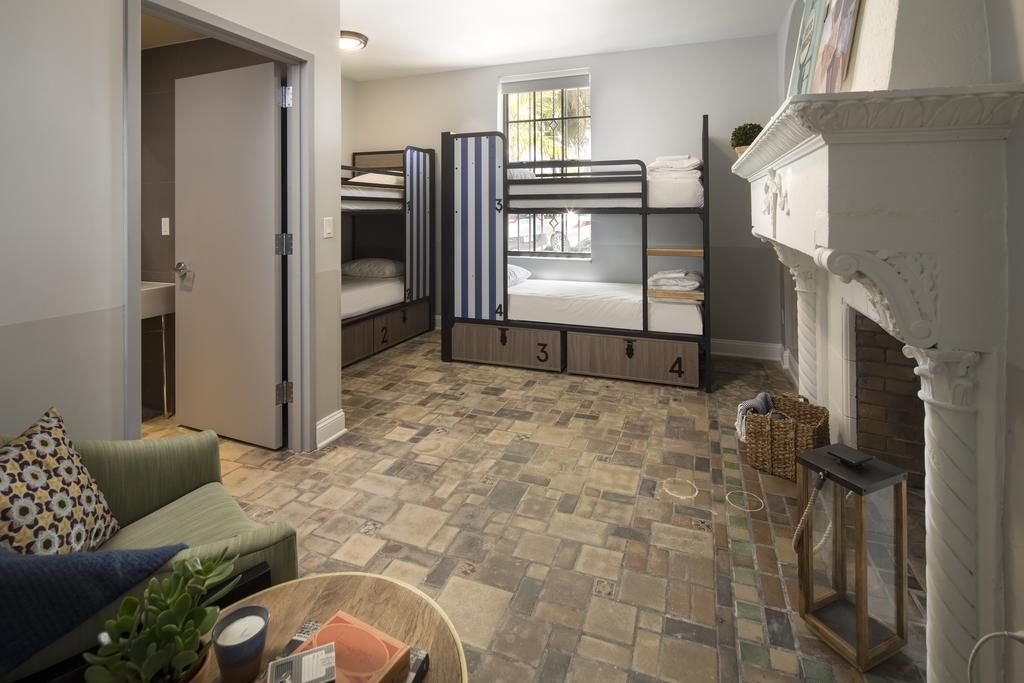 A imagem mostra um quarto com decoração acolhedora e piso de ladrilhos multicoloridos no Generator Miami. Há dois beliches com quatro camas no total, cada uma com um número na base e cortinas para privacidade. À esquerda, uma porta aberta revela um banheiro. Em primeiro plano, há uma poltrona verde com almofadas e uma mesa de centro de madeira com plantas e livros. À direita, uma lareira branca decorativa adiciona charme ao ambiente. A janela permite a entrada de luz natural, iluminando suavemente o espaço.