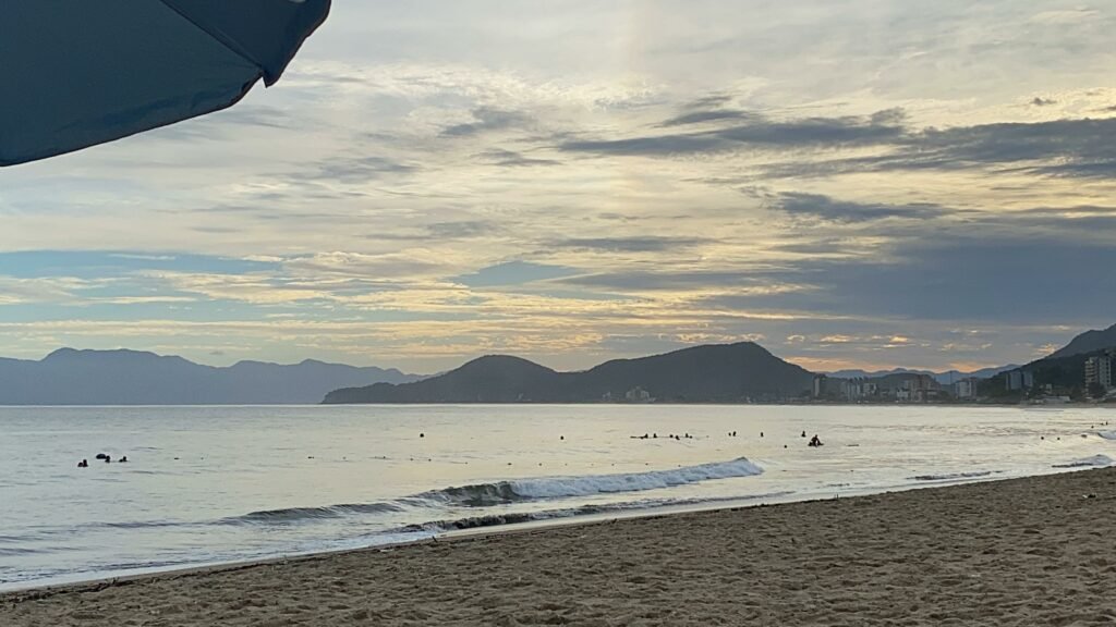Pôr do sol na Praia da Cocanha, em Caraguatatuba, no Litoral Norte de São Paulo. O mar calmo bate na areia da praia no fim da tarde, e ao fundo é possível ver algumas colinas da cidade.