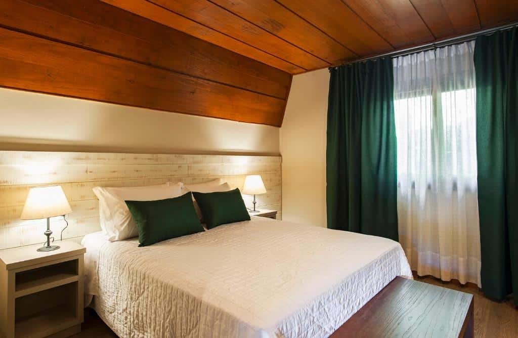 A foto mostra um quarto bem arrumado com uma cama grande no centro, com roupa de cama branca e dois travesseiros, além de um travesseiro decorativo verde. A cabeceira é de madeira clara, combinando com o teto de madeira. Em cada lado da cama, há mesas de cabeceira brancas com abajures em cima. Há uma janela na direita com cortinas verdes que permitem a entrada de luz natural, criando uma atmosfera acolhedora e convidativa.