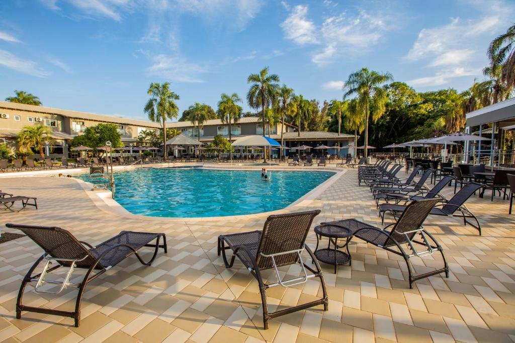 Parte do Grand Carimã Resort & Convention Center que mostra uma piscina ao ar livre com várias espreguiçadeiras em volta. Ao fundo tem parte da construção do resort e algumas palmeiras, imagem ilustrando post Resorts em Foz do Iguaçu.