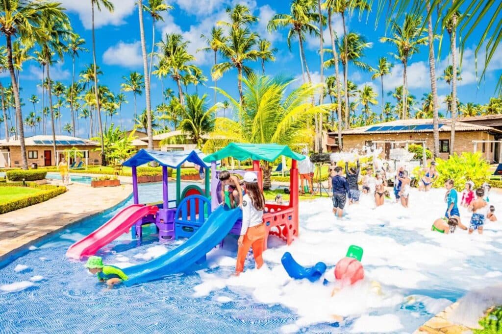 Crianças brincam em uma piscina com escorregadores coloridos e espuma do Grand Oca Maragogi All Inclusive Resort. Palmeiras e prédios estão ao fundo sob um céu azul brilhante.