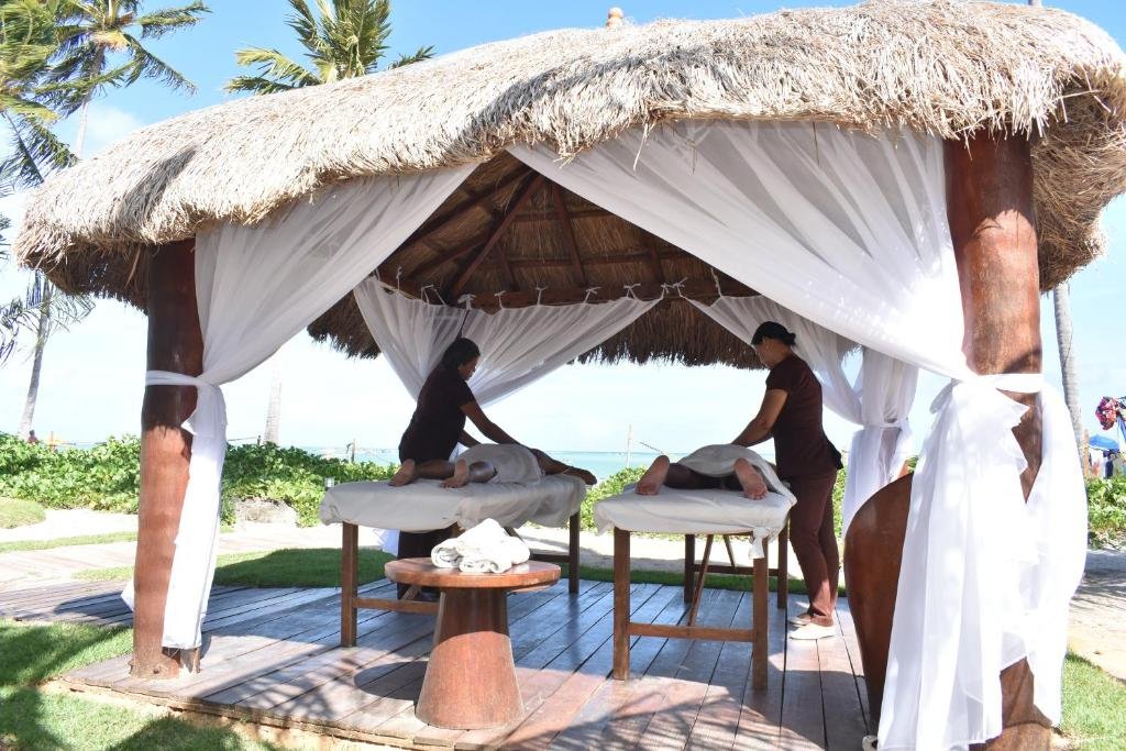 Duas pessoas recebem massagens em mesas sob um gazebo de palha na praia, cercado por cortinas brancas e palmeiras.