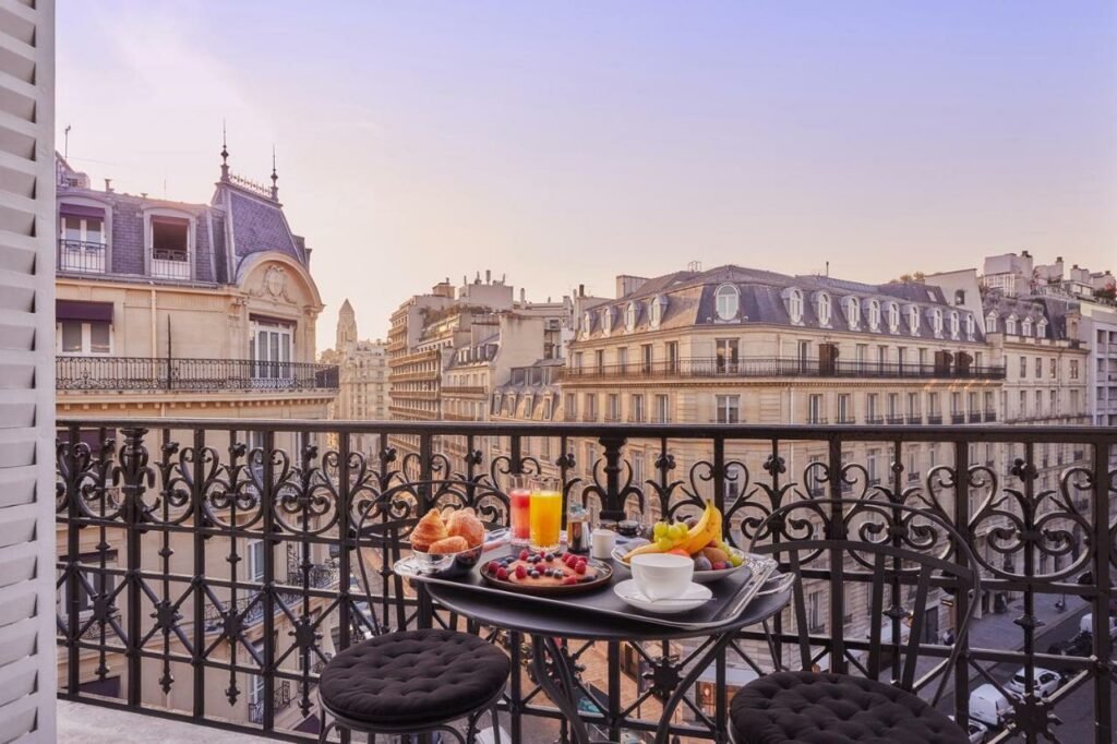 A imagem mostra uma mesa redonda em uma varanda do Grand Powers Hotel com vista para prédios históricos em uma cidade ao amanhecer. Na mesa, há croissants, frutas frescas, dois copos de suco, uma xícara de café e uma taça com frutas vermelhas. A varanda tem uma grade de ferro trabalhada e duas cadeiras pretas com assentos estofados. Ao fundo, o céu está em tons suaves de rosa e lilás, iluminando os edifícios ao redor.
