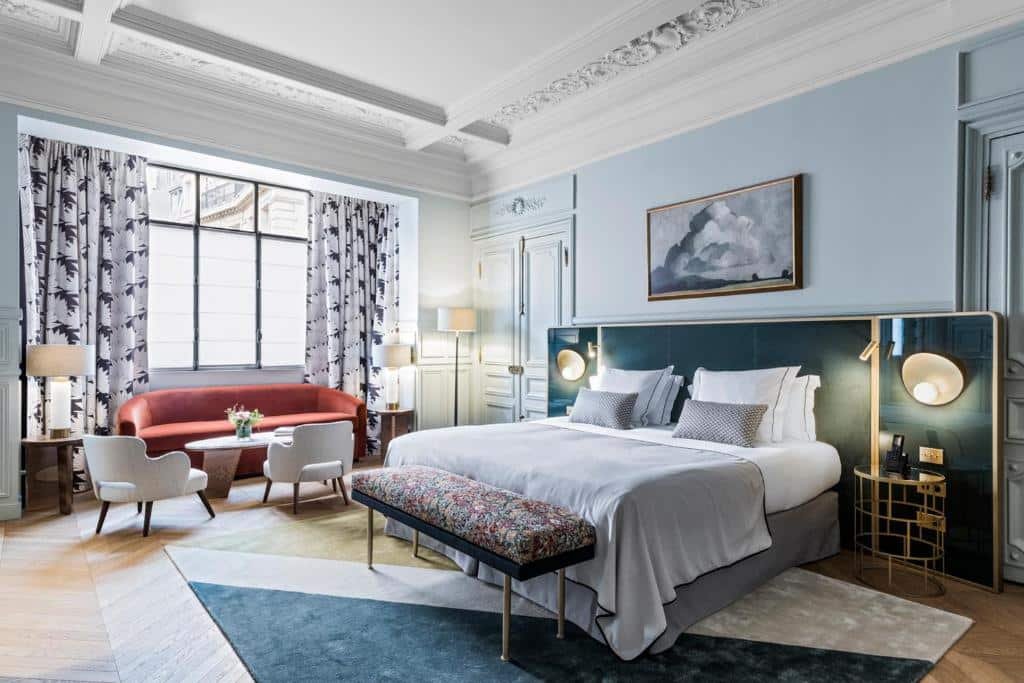 A imagem mostra um quarto luxuoso e bem iluminado no Grand Powers Hotel, um dos hotéis com café da manhã em Paris. A cama é grande, com lençóis brancos e várias almofadas decorativas, posicionada contra uma cabeceira azul escura com luminárias modernas. Há um banco estofado com padrão floral aos pés da cama. À direita, há uma grande janela com cortinas estampadas em preto e branco, e em frente à janela, um sofá vermelho e duas cadeiras brancas ao redor de uma mesa de centro. O teto tem detalhes ornamentados e a decoração é sofisticada, com tons suaves e móveis elegantes.