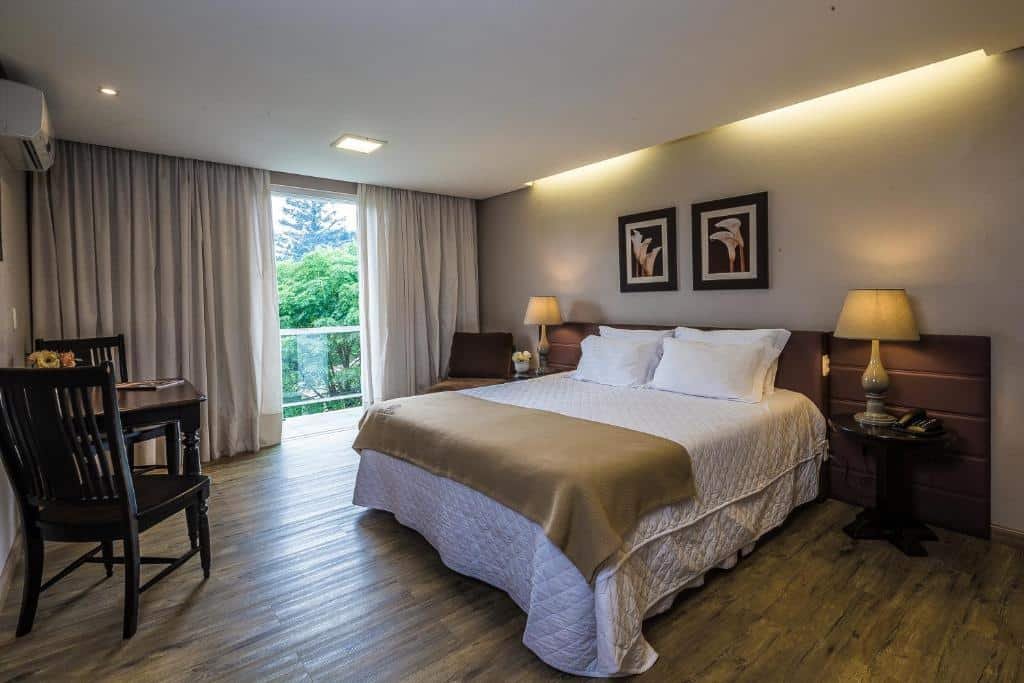Um quarto bastante espaçoso no Grande Hotel Canela. A cama é king-size, e está no centro da imagem, inclinada à direita. Atrás dela há uma cabeceira e madeira escura e uma mesa de cabeceira em seus dois lados com um abajur em cima. Uma grande janela com varanda está no lado esquerdo da cama. Na frente da cama, encostada na parede à esquerda, há uma mesa com duas cadeiras e um ar-condicionado fixo no topo do teto.