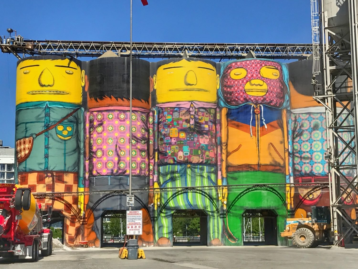 Cinco grandes silos pintados com figuras coloridas e caricaturais em vários padrões e cores, localizados em uma área industrial ao ar livre. Imagem para ilustrar post sobre o que fazer em Vancouver.