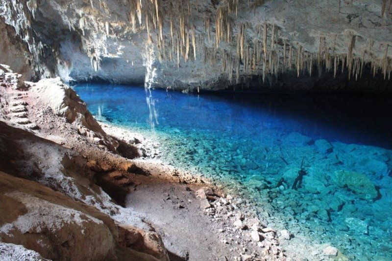 Interior da caverna com um lago subterrâneo azul claro, estalactites penduradas no teto e uma costa rochosa que leva à água.