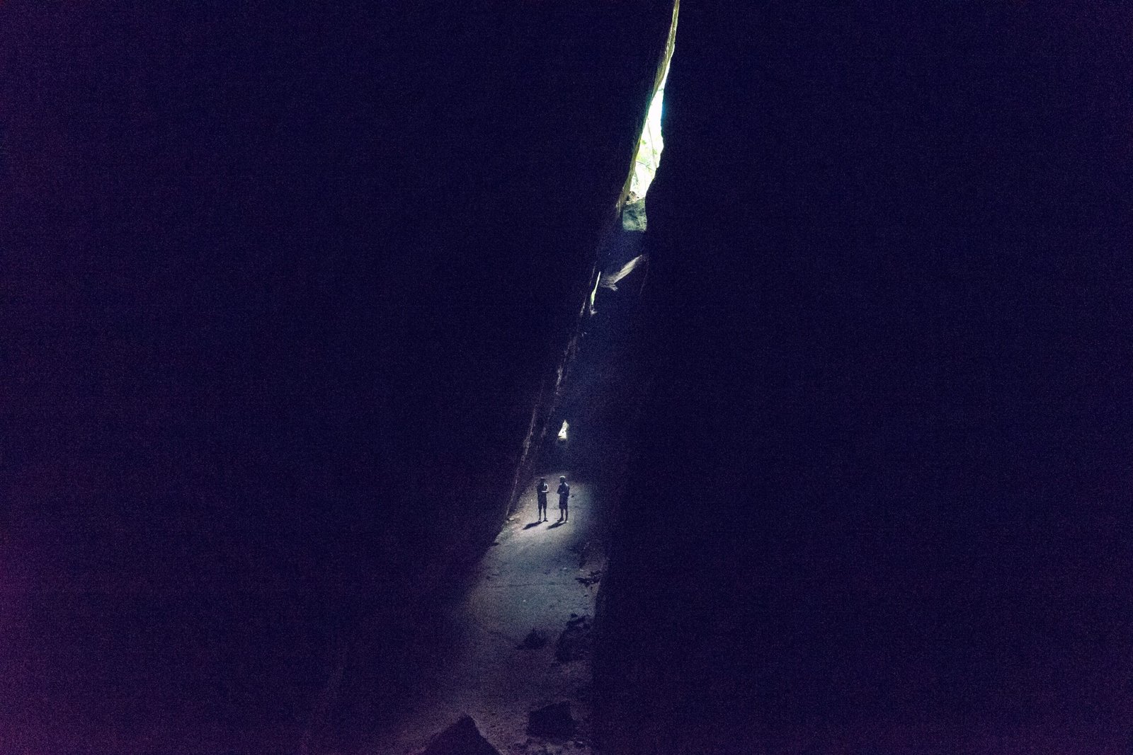 Duas pessoas estão em uma caverna estreita e escura com luz vindo de cima, projetando sombras no chão rochoso.