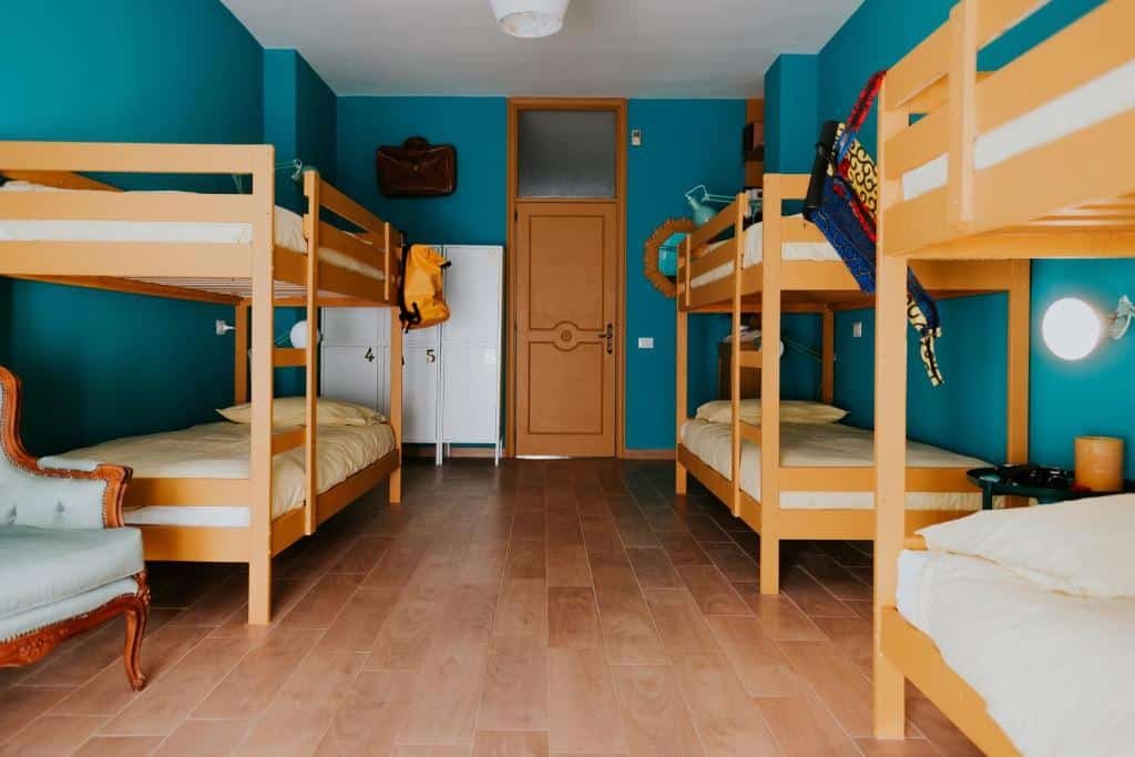 Quarto do hostel Habari We Dorm com três beliches, um do lado esquerdo da foto e dois do lado direito. As paredes do quarto são azuis, tem uma poltrona do lado esquerdo e ao fundo tem um armário com três portas e ao lado uma porta de madeira. Ilustrando post Hotéis em Puglia.