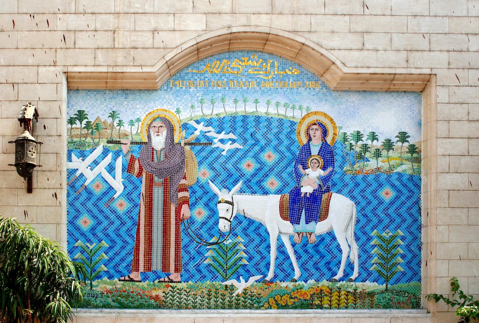 Um mosaico colorido retrata três figuras religiosas: uma de pé com um cajado e duas montadas em um burro, contra um fundo azul padronizado no Hanging Church.