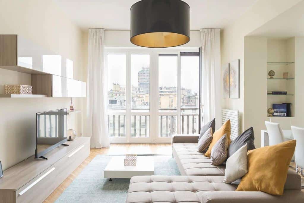 Sala ampla do Heart Milan Apartments San Sepolcro Duomo com um sofá com diversas almofadas, um abajur, uma varanda ampla com cortinas, há um rack com uma televisão e uma pequena mesa de centro, em Milão