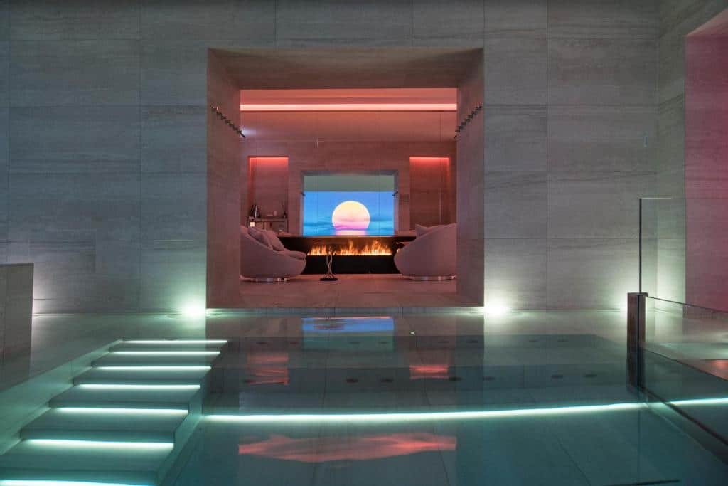 Spa do Helvetia&Bristol Firenze, um dos hotéis em Florença, com hidromassagem iluminada e uma área com duas poltronas brancas redondas e um telão mostrando imagem de fogo (imitando uma lareira) e um pôr do sol rosado