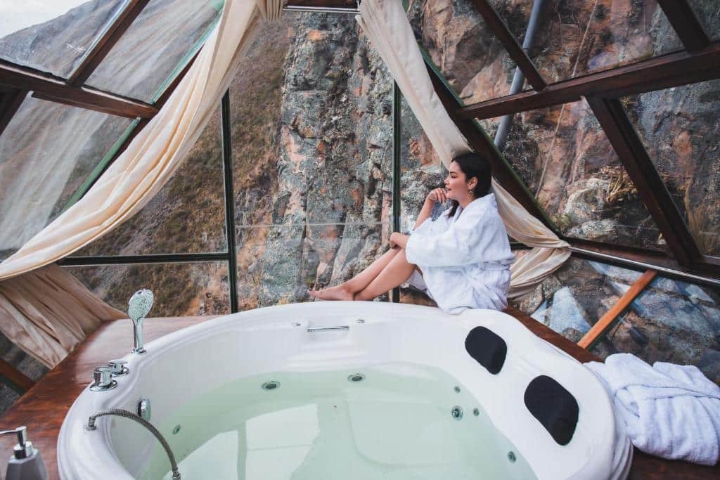 área de jacuzzi do Vertical Sky Luxury Suites com uma mulher branca, vestida com um roupão atoalhado, sentada ao lado da jacuzzi, olhando para a paisagem de vidro.