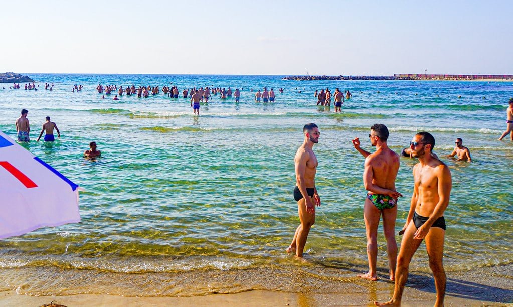 Pessoas vadeando e nadando na praia Hilton em Tel Aviv. Três homens em trajes de banho conversam perto da costa. O oceano se estende ao fundo.