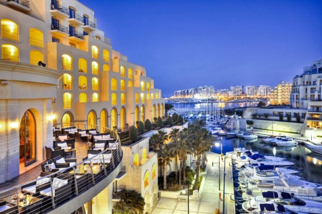 Vista das sacadas do Hilton Malta de frente para um deck com muitos barcos e lanchas, a varanda do hotel está com diversos sofás e, na entrada do local, há diversos coqueiros