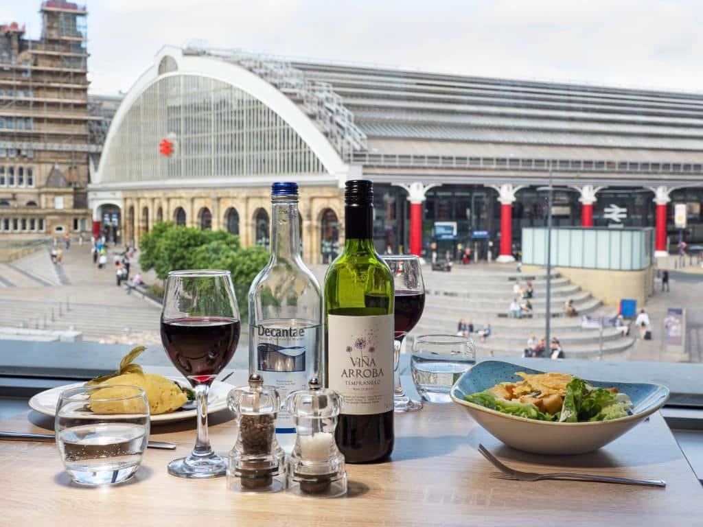 Mesa com taça e garrafa de vinho, além de dois pratos com comida e potes com temperos, com janela de vidro ao lado e vista de uma praça que fica em frente ao Holiday Inn Liverpool City Centre, an IHG Hotel