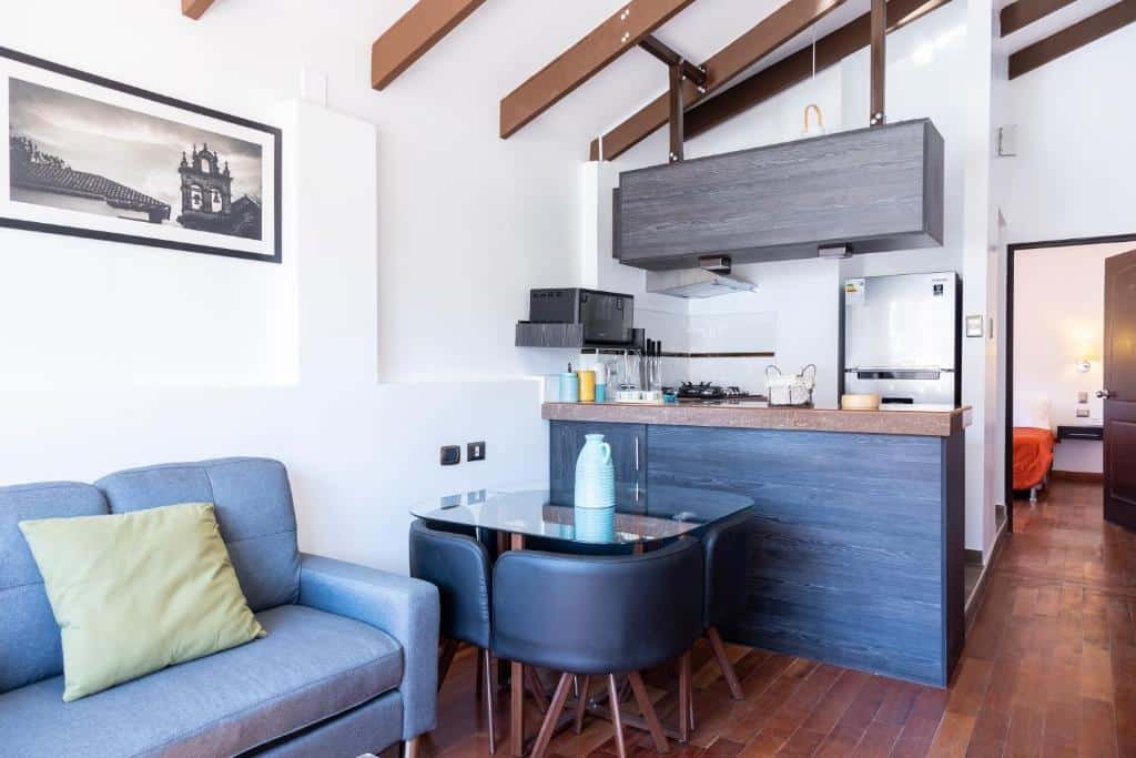 apartamento do Hostal & Apartments El Triunfo com uma sofá azul no canto inferior esquerdo, ao lado de uma pequena mesa de vidro com quatro lugares, em frente ao balcão de uma área aberta e reservada para a cozinha. Ao fundo, no canto direito, há uma porta aberta com vista para o quarto.