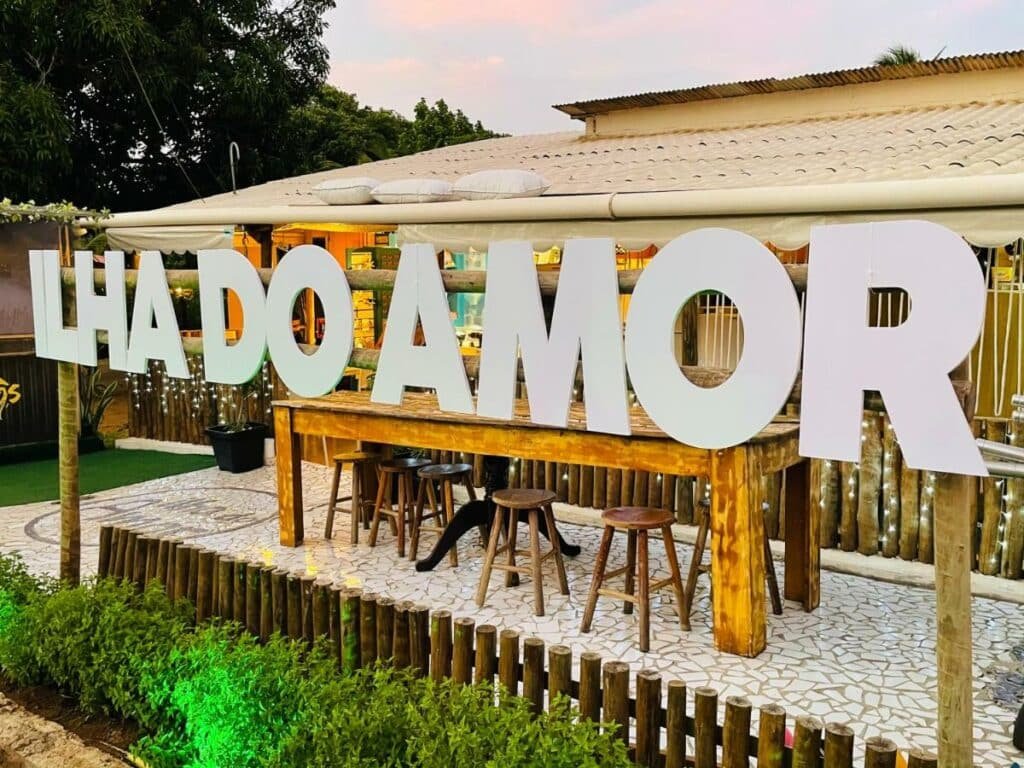 Letreiro branco escrito "ILHA DO AMOR", em área externa do hostel de mesmo nome, com uma mesa grande e banquinhos atrás