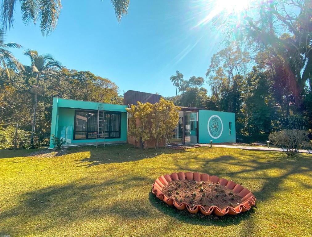 Casa verde do Hostel Jardim de Lótus, com portas e janelas de vidro grandes transparente, um gramado na frente com um jardim redondo ao meio e várias árvores ao redor. No céu há um raio de sol iluminando o céu azul