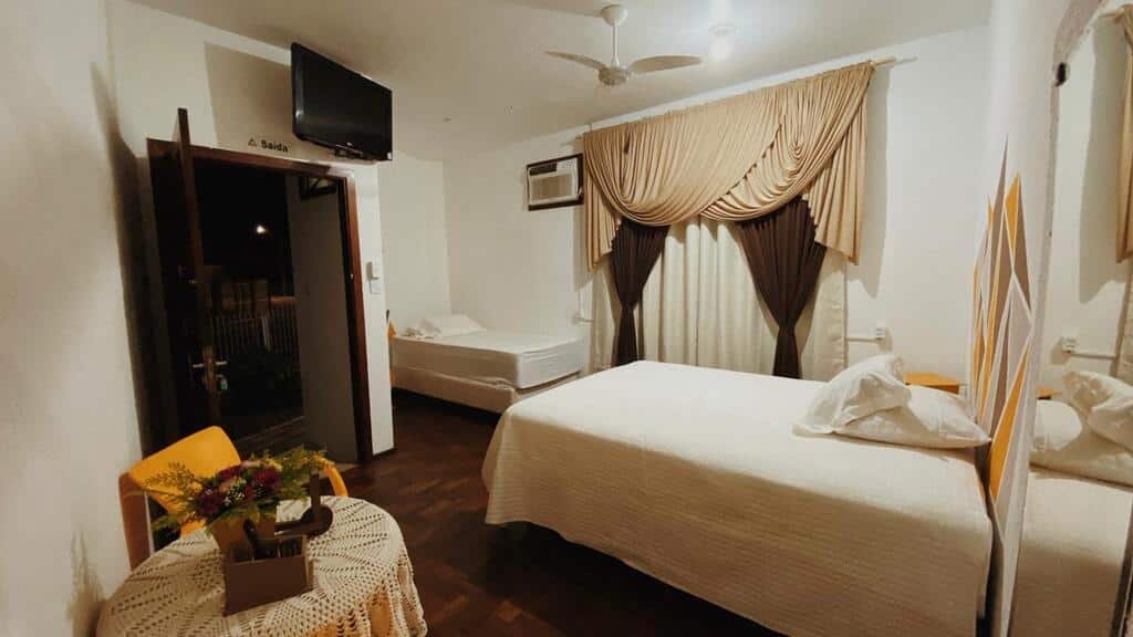 Quarto do Hostel Lumaria, com uma cama de casal, e uma de solteiro, uma cortina com três camadas em diferentes cores, sendo uma marrom, uma bege e outra creme, um ventilador, ar-condicionado, TV, mesa redonda com uma cadeira amarela e uma porta mostrando o lado de fora da pousada durante uma noite