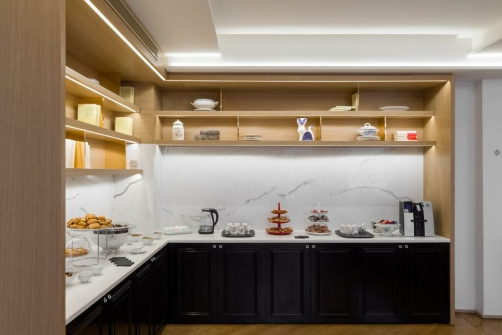 A imagem mostra uma área de café da manhã moderna e organizada no Hotel 34B - Astotel. Há um balcão branco com armários pretos abaixo e prateleiras de madeira acima, decoradas com livros, louças e enfeites. Sobre o balcão, há bandejas de croissants, doces variados, uma jarra elétrica, uma cafeteira, xícaras e tigelas. A parede ao fundo tem acabamento em mármore branco com veios cinza, e a iluminação é suave, criando um ambiente acolhedor e sofisticado.