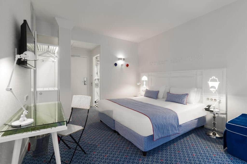 A imagem mostra um quarto moderno e bem iluminado no Hotel 34B - Astotel, um dos hotéis com café da manhã em Paris. A cama grande tem roupa de cama branca com detalhes em azul e travesseiros combinando. As paredes são brancas, com luminárias modernas ao lado da cama. Há uma mesa de vidro com uma cadeira branca ao lado, e uma televisão montada na parede acima. O chão tem carpete azul com pequenos detalhes em vermelho. No fundo, há um armário aberto e uma porta que leva ao banheiro, criando um ambiente organizado e aconchegante.