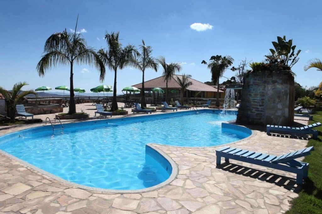 Área de piscina ao ar livre com palmeiras, espreguiçadeiras, guarda-sóis verdes e uma passarela de pedra sob um céu azul claro no Hotel Alpha Ville Chalés, um dos hotéis fazenda em Minas Gerais.