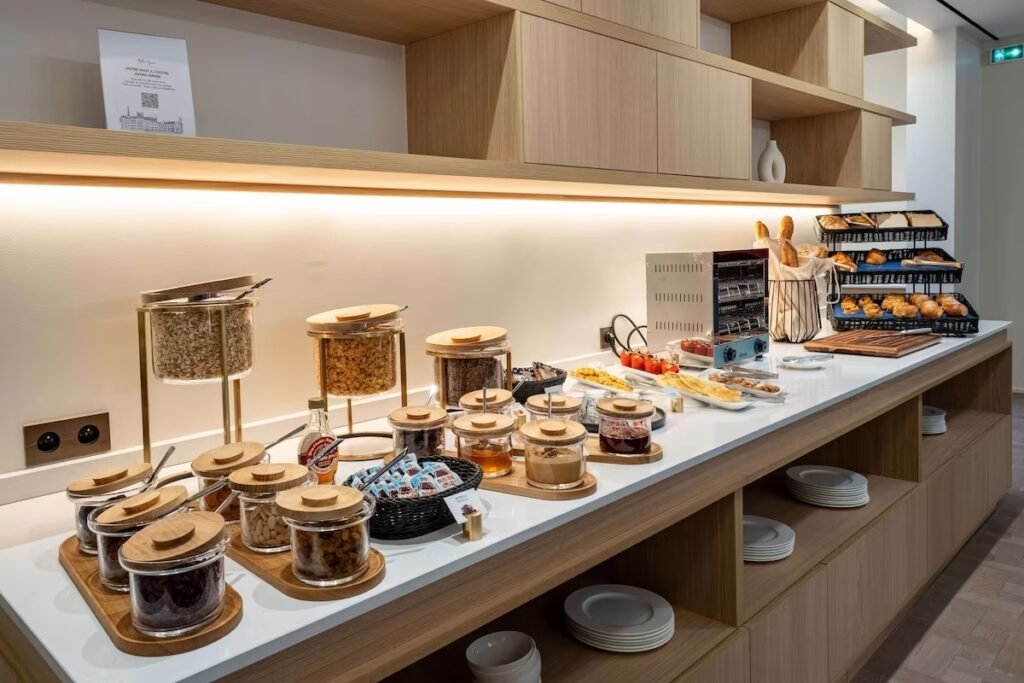 A imagem mostra um buffet de café da manhã bem organizado em um balcão de madeira clara no Hotel Astra Opera - Astotel. Há diversos potes de vidro com tampas de madeira, contendo cereais, frutas secas, nozes e granolas. Ao lado, há cestas com pães, baguetes e croissants frescos, além de uma torradeira elétrica. Há também potes de geleia, mel e outros condimentos, e pratos brancos organizados nas prateleiras inferiores. O ambiente é iluminado de forma suave, criando uma atmosfera acolhedora e funcional.