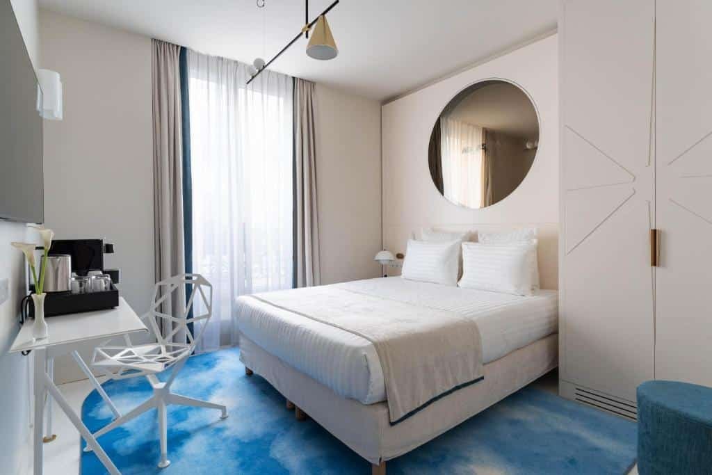 A imagem mostra um quarto moderno e bem iluminado no Hotel Astra Opera - Astotel, um dos hotéis com café da manhã em Paris. A cama é grande, com roupa de cama branca e dois travesseiros, posicionada contra uma parede clara com um grande espelho redondo acima da cabeceira. O quarto possui uma janela alta com cortinas translúcidas, permitindo a entrada de luz natural. Há um tapete azul claro no chão, uma cadeira branca de design geométrico e uma mesa com um conjunto de café. A decoração é minimalista e elegante, criando um ambiente tranquilo e acolhedor.