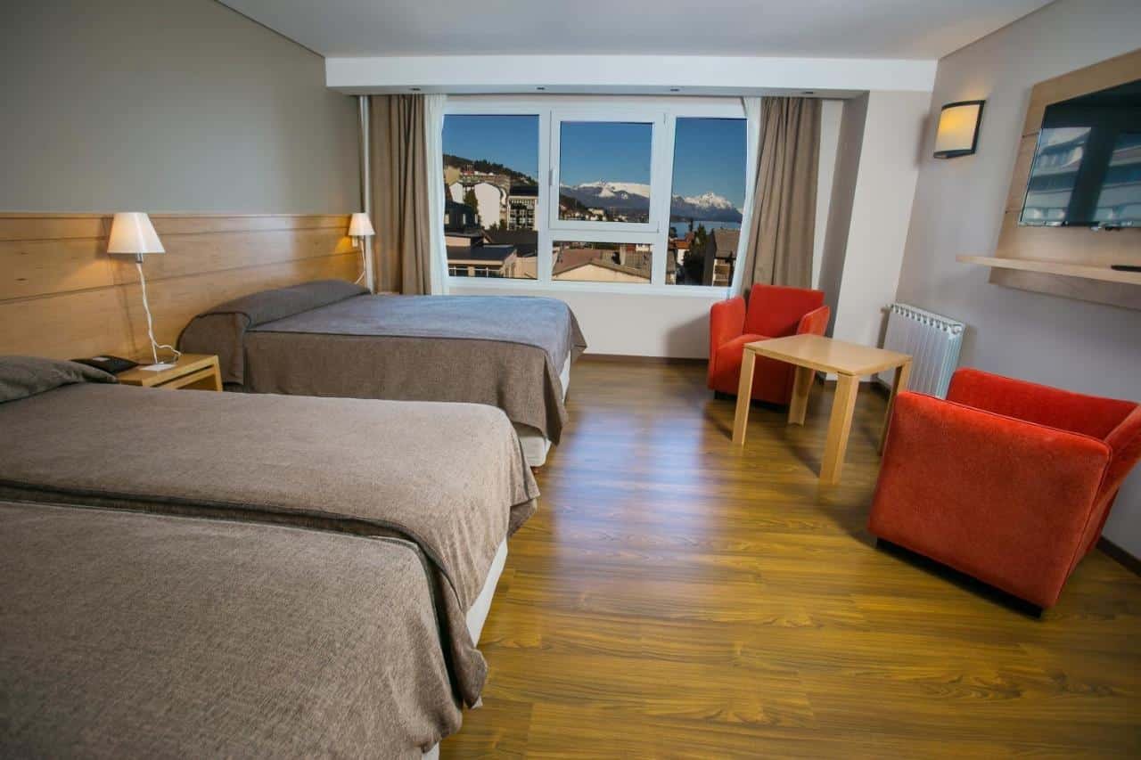 Quarto do Hotel Ayres Del Nahuel. Duas camas de solteiro e uma cama de casal, do lado direito duas poltronas, uma mesa de centro e uma TV. No fundo uma janela de vidro.