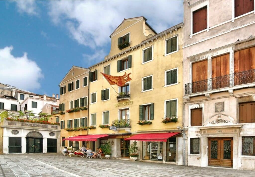 Fachada do Hotel Bel Sito e Berlino em uma construção antiga em uma praça bem típica, com janelas verdes com flores e, na entrada, um toldo vermelho com cadeiras e mesas embaixo