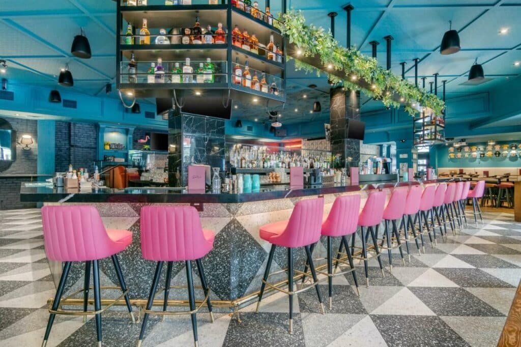 Bar do Hotel Belmont Vancouver MGallery com um balcão de marm´óre extenso com diversas banquetas rosas pink para se sentar, o chão é cinza e branco, no teto há diversos lustres