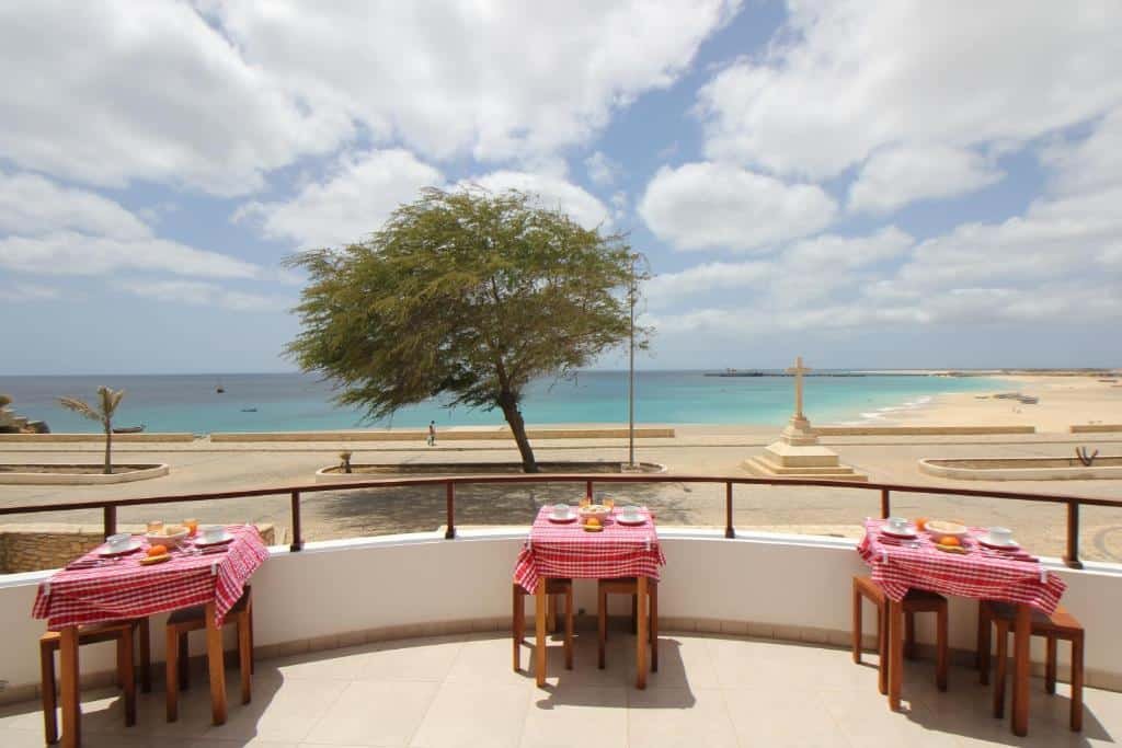 Parte do Hotel Casa Evora na Vila do Maio que mostra uma varanda aberta com três mesas e cadeiras com vista para a rua com árvores e para o mar azul. Imagem ilustrando post Hotéis em Cabo Verde.