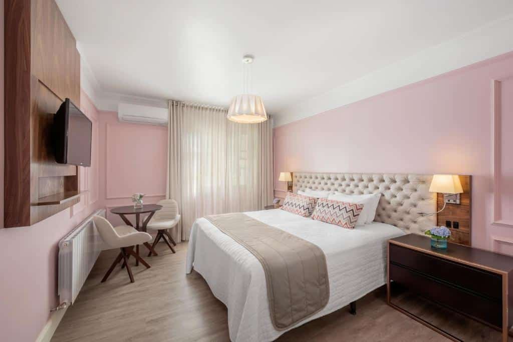 Quarto com paredes rosa claro no Hotel Casacurta, com cabeceira de botonê, luzes de leitura em cada lado, mesa de cabeceira com flores, TV na parede, janela com cortinas fechadas, aquecedor, mesa com duas cadeiras e ar-condicionado