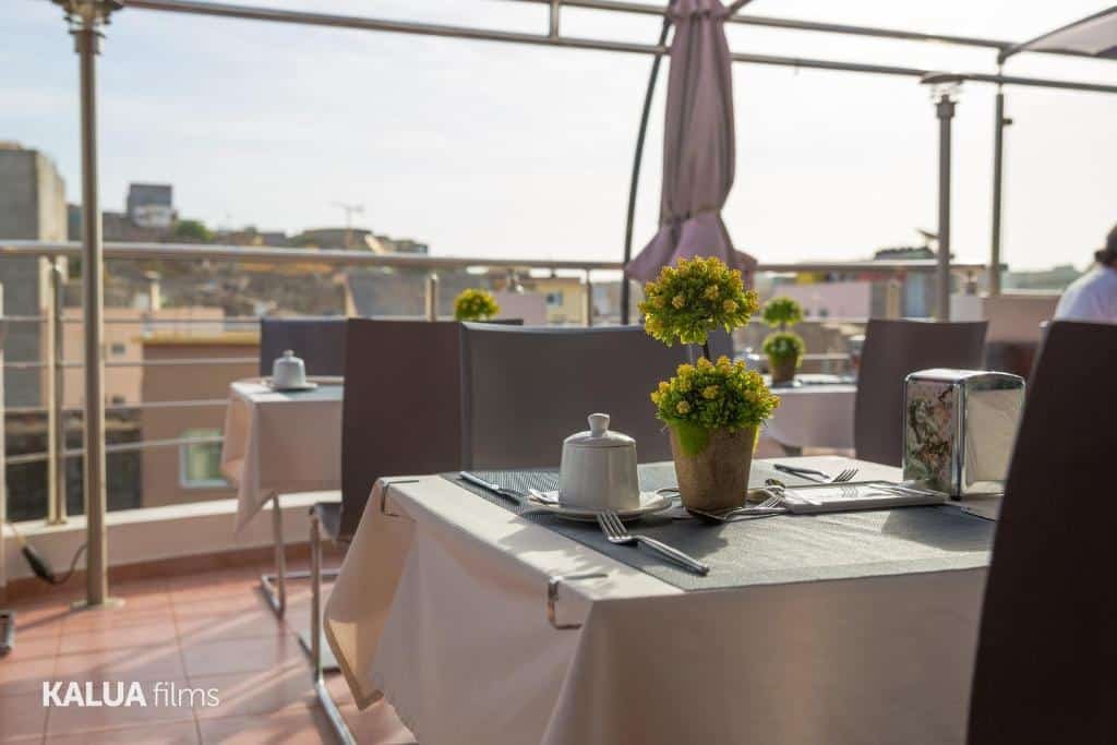 Parte do Hotel Cesaria em Praia que mostra em primeiro plano uma mesa quadrada com alguns objetos em cima e duas cadeiras. Em volta dessa mesa tem outras parecidas. Esse espaço é aberto e tem vista para as construções da cidade. Imagem ilustrando post Hotéis em Cabo Verde.