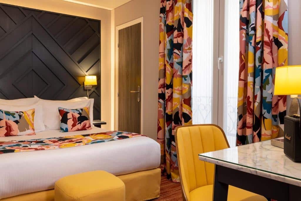 Quarto do Hôtel des Arts Montmartre, um dos hotéis com café da manhã em Paris. Uma cama de casal está encostada na parede ao lado esquerdo com mesinhas de cabeceira e luminárias aos dois lados, assim como um banco aos pés. Ao lado fica uma janela com cortinas, e em frente à cama há uma mesa com cadeira e abajur.
