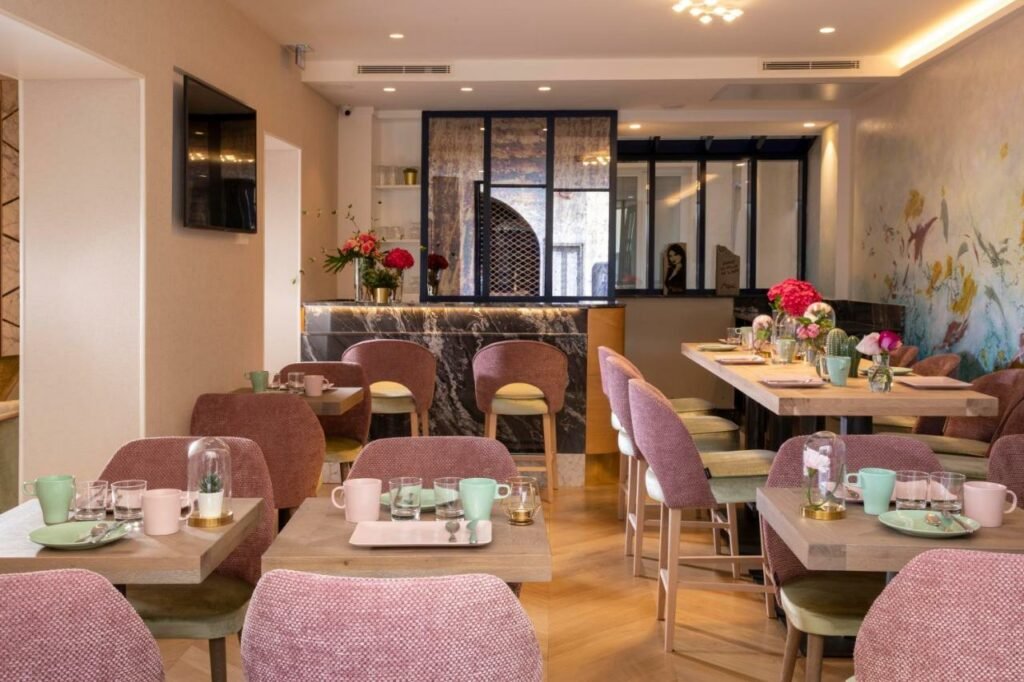 Salão do Hôtel des Arts Montmartre com cadeiras e mesas postas para o café. Há vasinhos de flores pelas mesas também, e uma TV na parede ao lado esquerdo.