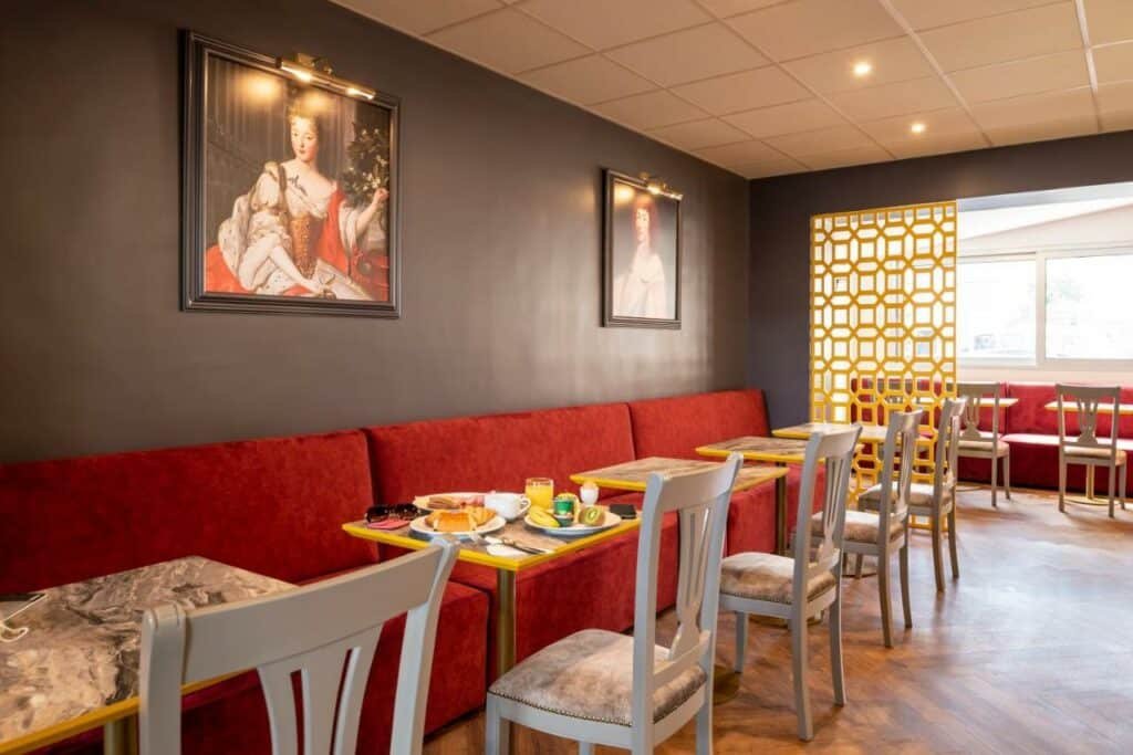 Sala de refeições do Hotel des Lys com sofás, mesinhas e cadeiras. Na parede acima do sofá há dois quadros. Na lateral direita um cobogó amarelo separa mais um local de refeições com janelas.
